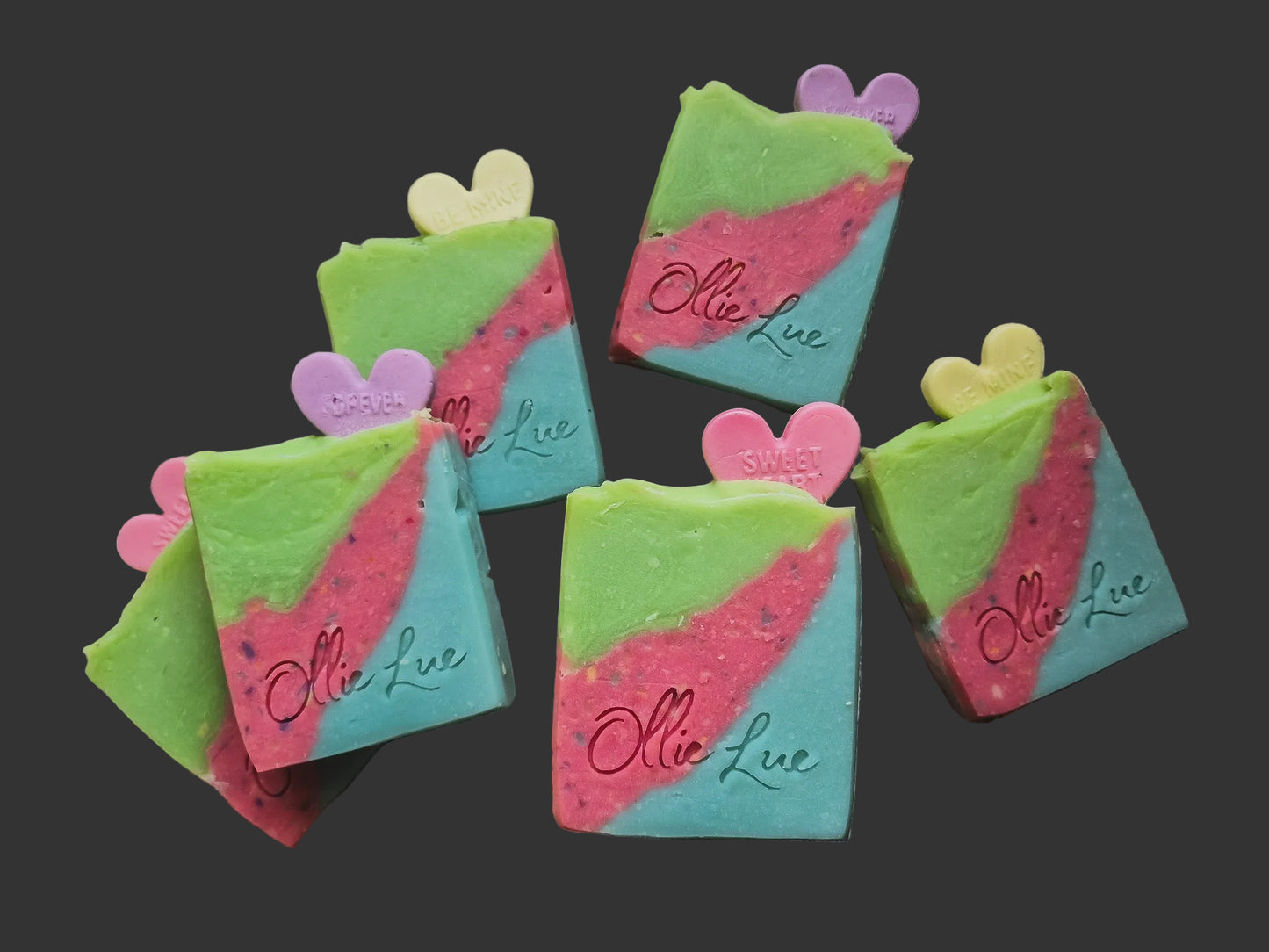 colorful layered soap stamped 'ollie lue' with pink, purple or yellow heart on top. the soap is layered green, pink, and blue all on a dark background