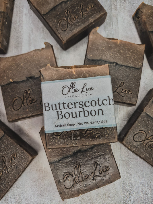 Several rustic rectangular bars of soap are arranged on a light textured surface, with one central bar wrapped in a paper label that reads 'Ollie Lue Soap Co. Butterscotch Bourbon.