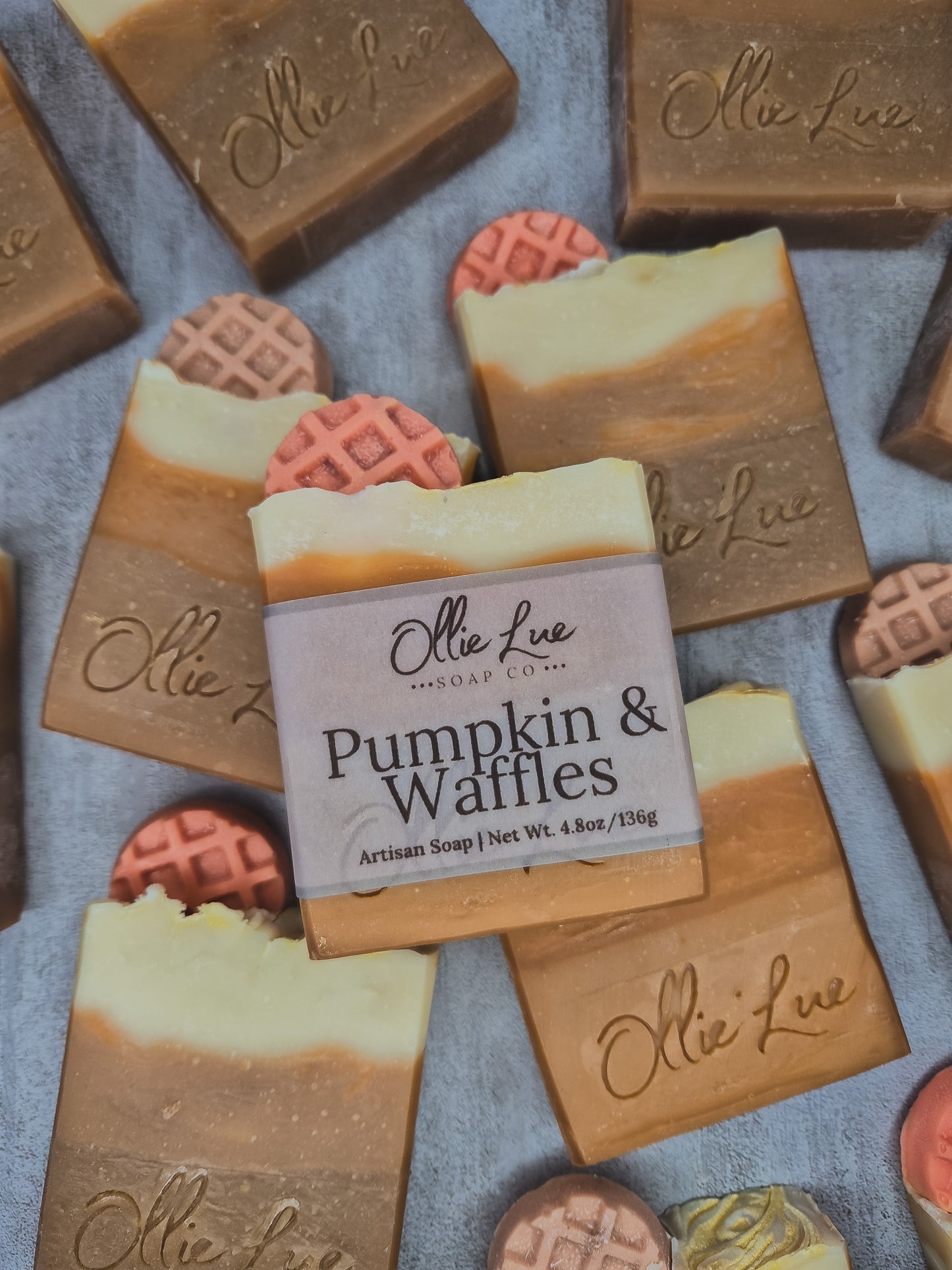 A bar of Ollie Lue 'Pumpkin & Waffles' artisan soap is centered, surrounded by other decorated soap bars on a textured gray background.