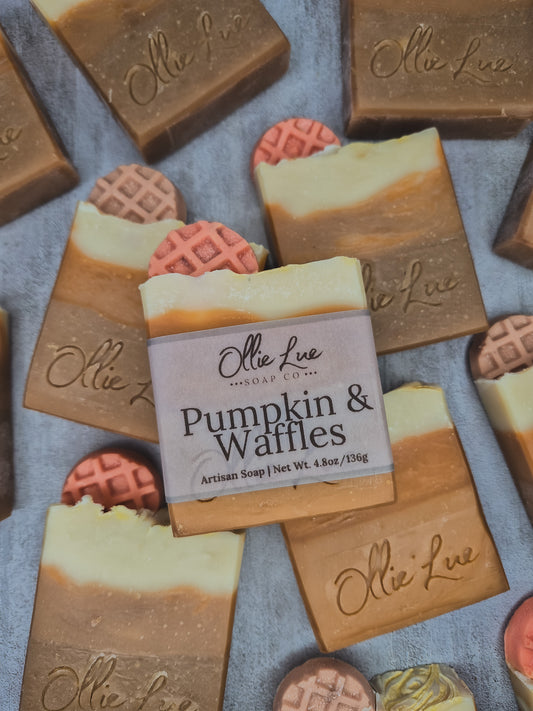 A bar of Ollie Lue 'Pumpkin & Waffles' artisan soap is centered, surrounded by other decorated soap bars on a textured gray background.