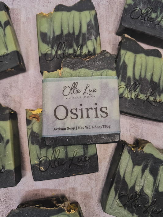 A bar of Osiris artisan soap by Olie Lue Soap Co. is centered on a textured light pink background, surrounded by several other marbled soap bars.