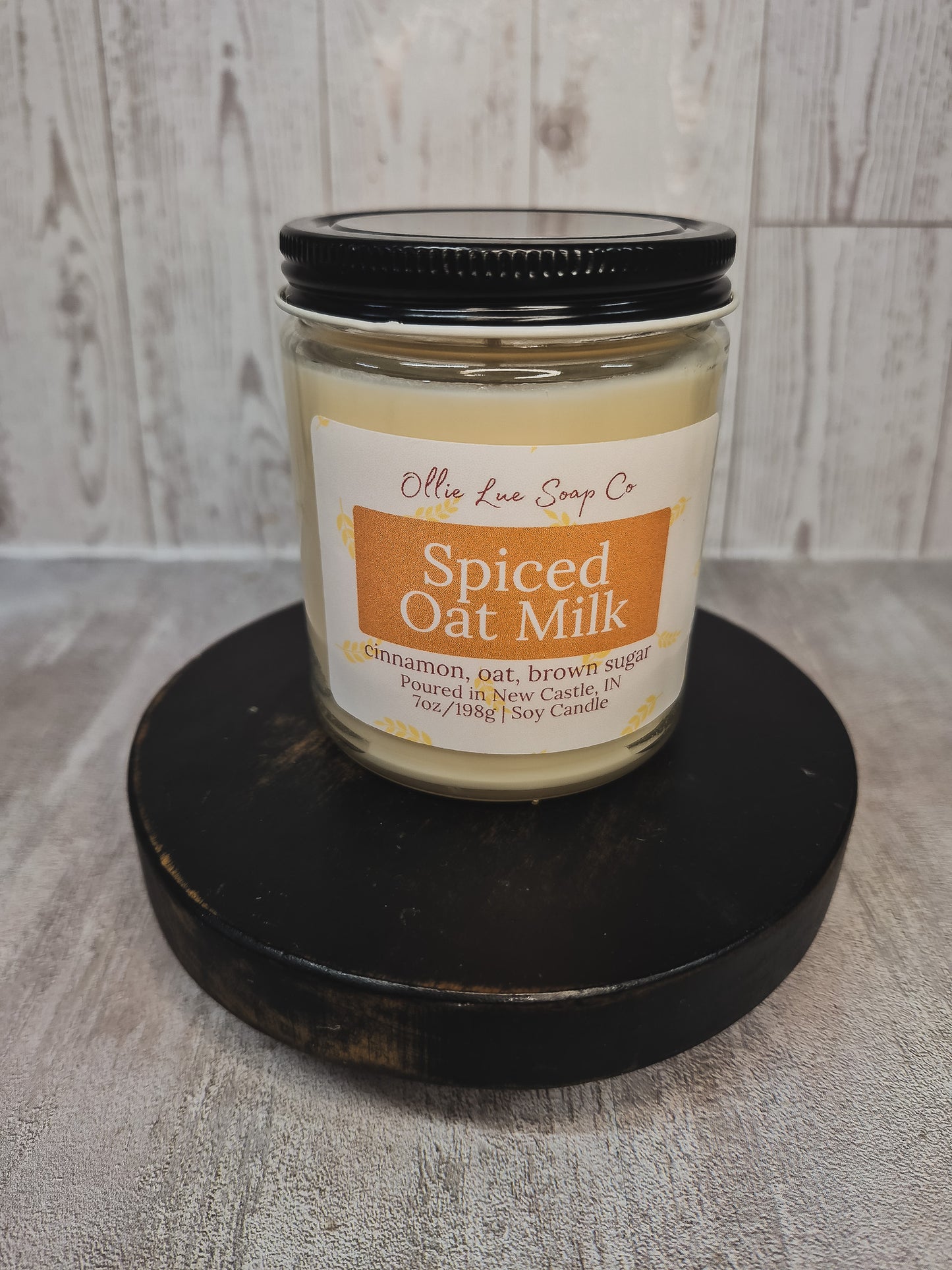 A jar candle labeled 'Spiced Oat Milk' by Ollie Lue Soap Co, sitting on a round black wooden platform against a light gray wood-plank background.