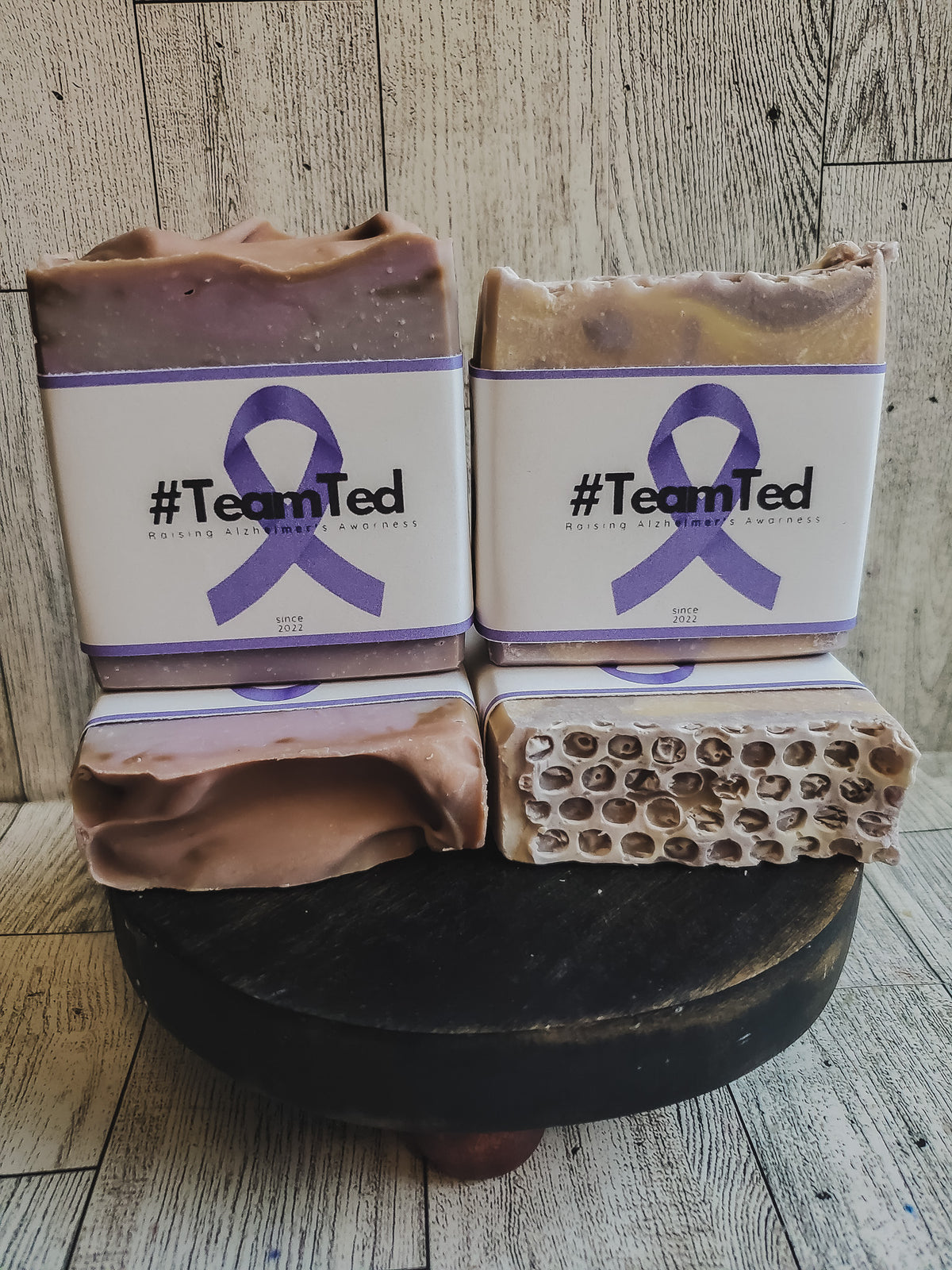 Two handmade soap bars with white labels featuring a purple ribbon and the text '#TeamTed' are displayed on a dark wooden stand against a rustic wood plank background. One soap bar is shown sliced to reveal a honeycomb-like internal structure.