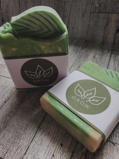 Two green and cream handmade soap bars wrapped in white paper bands with a circular green leaf logo that reads 'GROW.' on a rustic gray wood background.