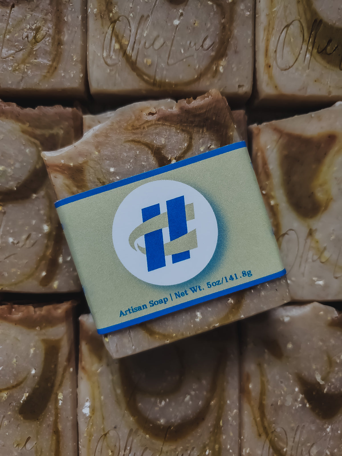A close-up of a handmade artisan soap bar wrapped in a light sage green paper band with a blue and white logo, set against a background of more textured soap bars.