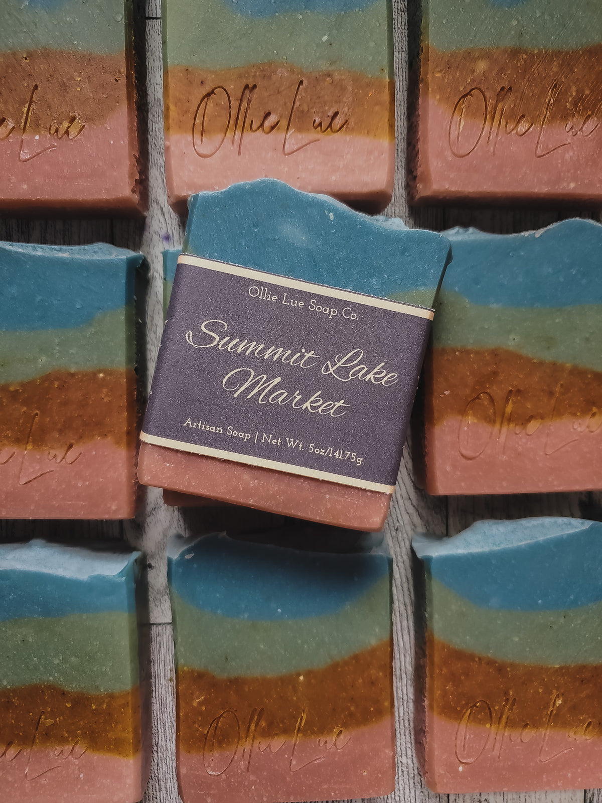 A close-up of handmade layered soap bars arranged in a grid. The center soap is labeled 'Summit Lake Market' by Ollie Lue Soap Co.