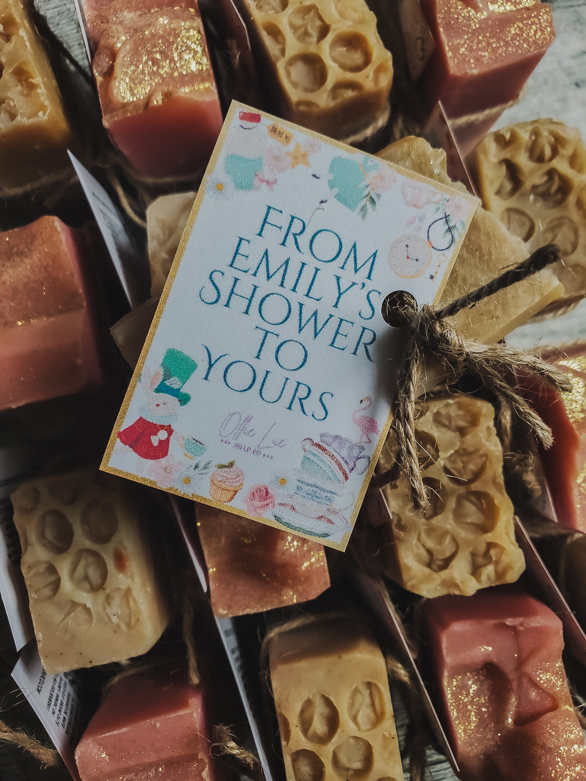 A gift box filled with handmade soap bars in pink, beige, and gold tones, with a central printed tag reading 'FROM EMILY'S SHOWER TO YOURS' tied with twine.