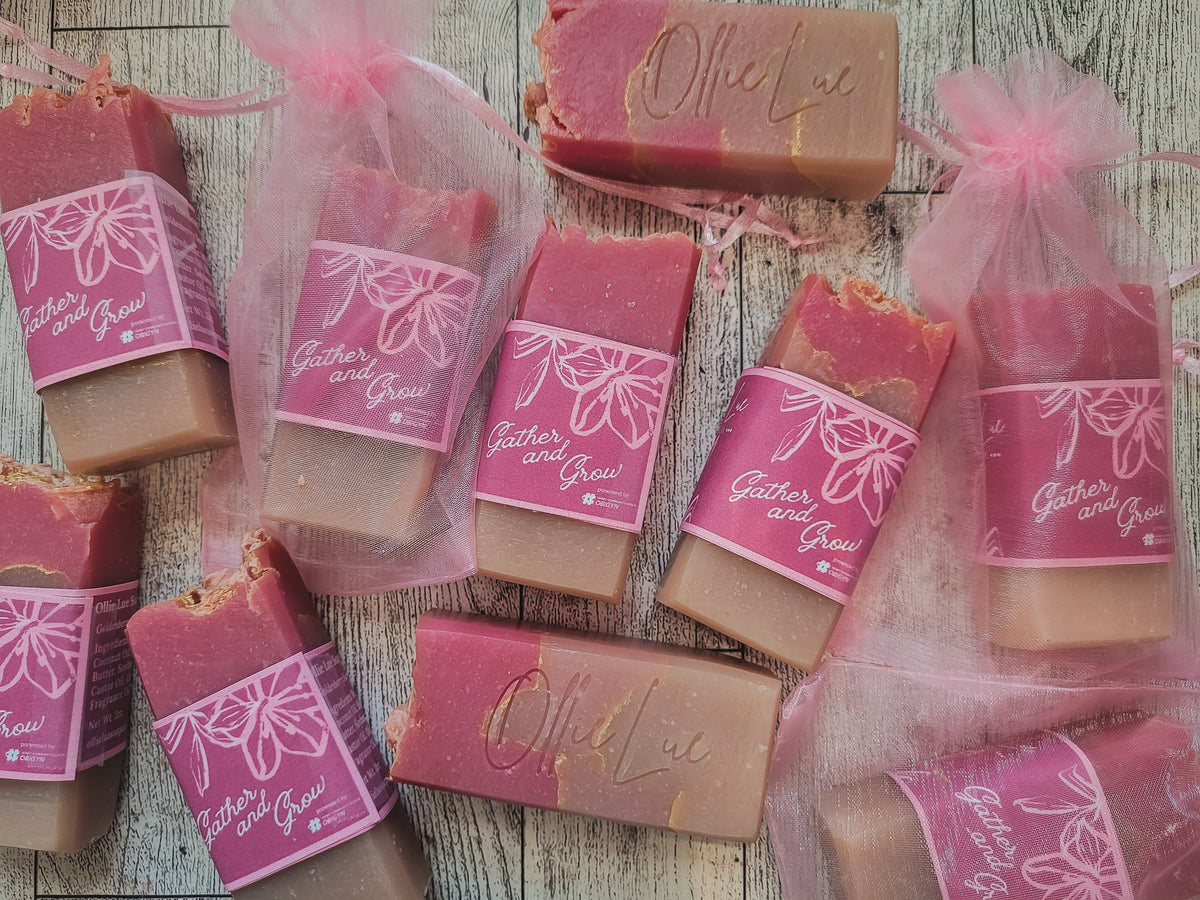 Several bars of handmade soap in pink and tan tones, some wrapped in sheer pink organza bags with pink ribbon, arranged on a rustic whitewashed wood surface. The labels read 'Gather and Grow' and one bar is embossed with 'Ollie Lue'.
