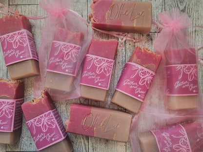 Several bars of handmade soap in pink and tan tones, some wrapped in sheer pink organza bags with pink ribbon, arranged on a rustic whitewashed wood surface. The labels read 'Gather and Grow' and one bar is embossed with 'Ollie Lue'.