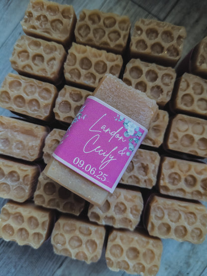 A close-up of multiple rectangular honeycomb-patterned soap bars arranged on a light wood surface, with one central bar wrapped in a bright pink paper label that reads 'Landon & Cassidy 09.06.