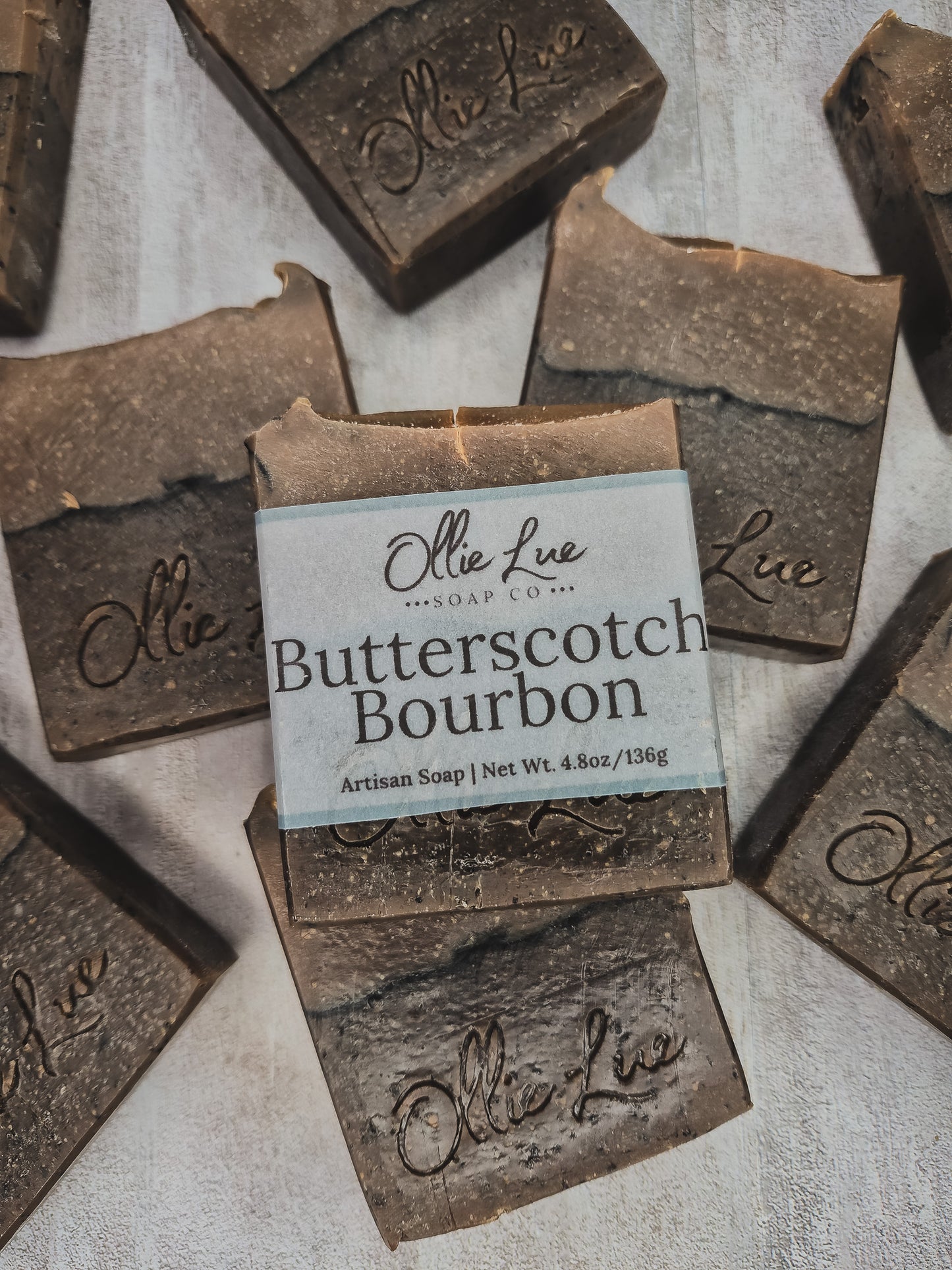 Several rustic rectangular bars of soap are arranged on a light textured surface, with one central bar wrapped in a paper label that reads 'Ollie Lue Soap Co. Butterscotch Bourbon.
