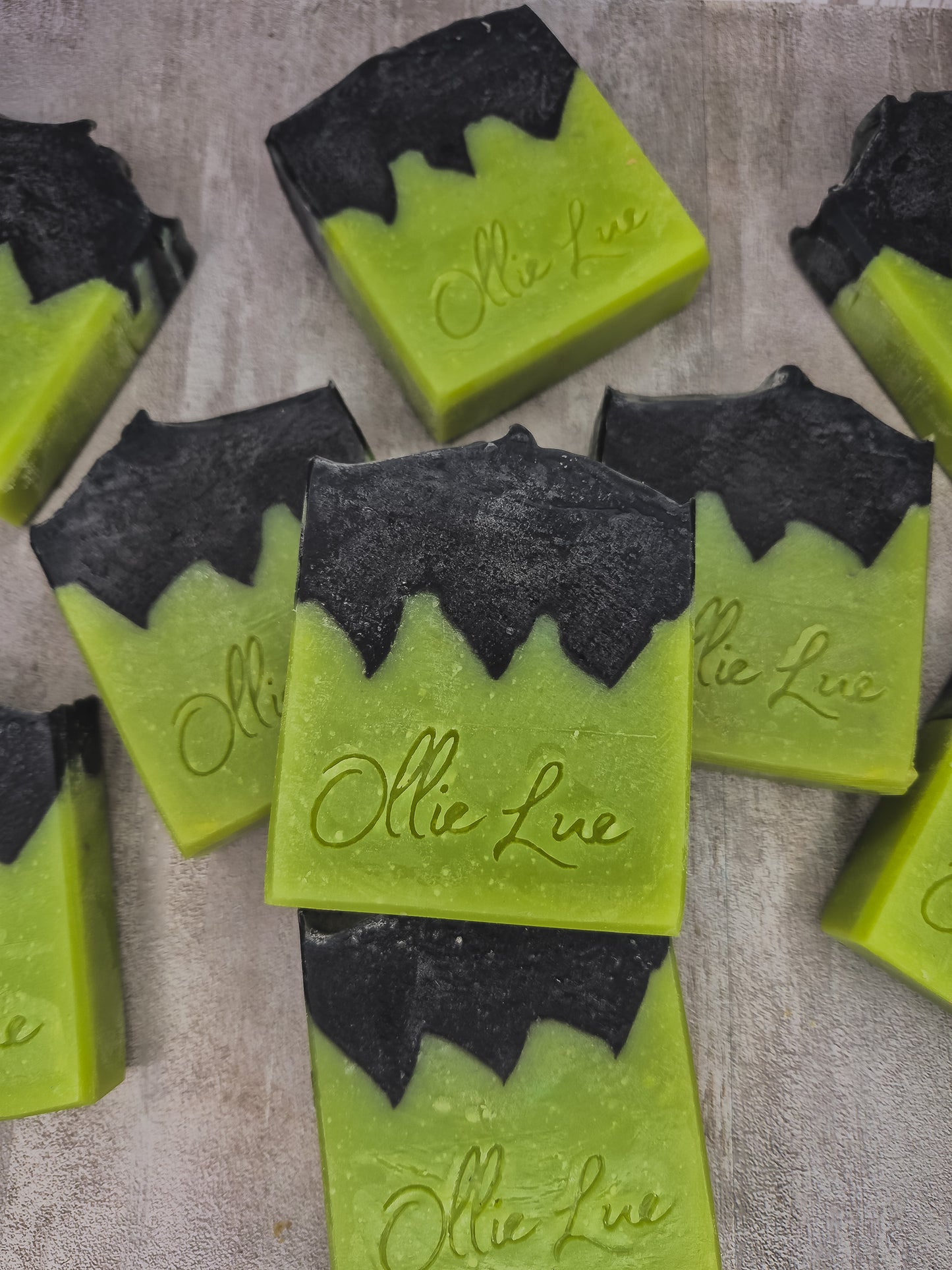 Several handmade rectangular bars of soap in bright lime green with black wavy top sections, each embossed with the name 'Ollie Lue, arranged on a light textured surface.