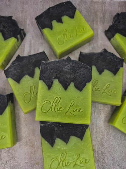 Several handmade rectangular bars of soap in bright lime green with black wavy top sections, each embossed with the name 'Ollie Lue, arranged on a light textured surface.