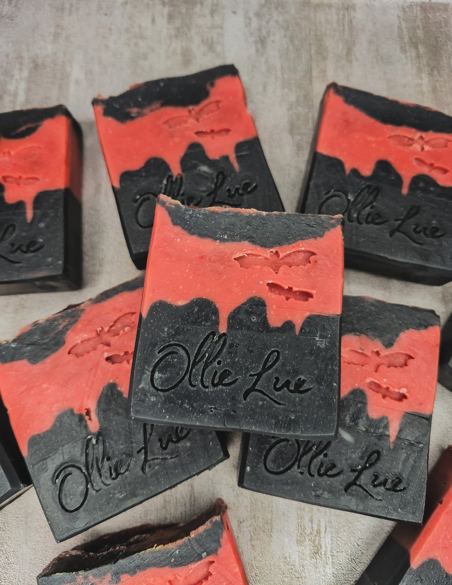 Several rectangular bars of handmade soap with a black and red marbled design, featuring a bat motif and the brand name 'Ollie Lue' embossed on the front, arranged on a neutral textured surface.