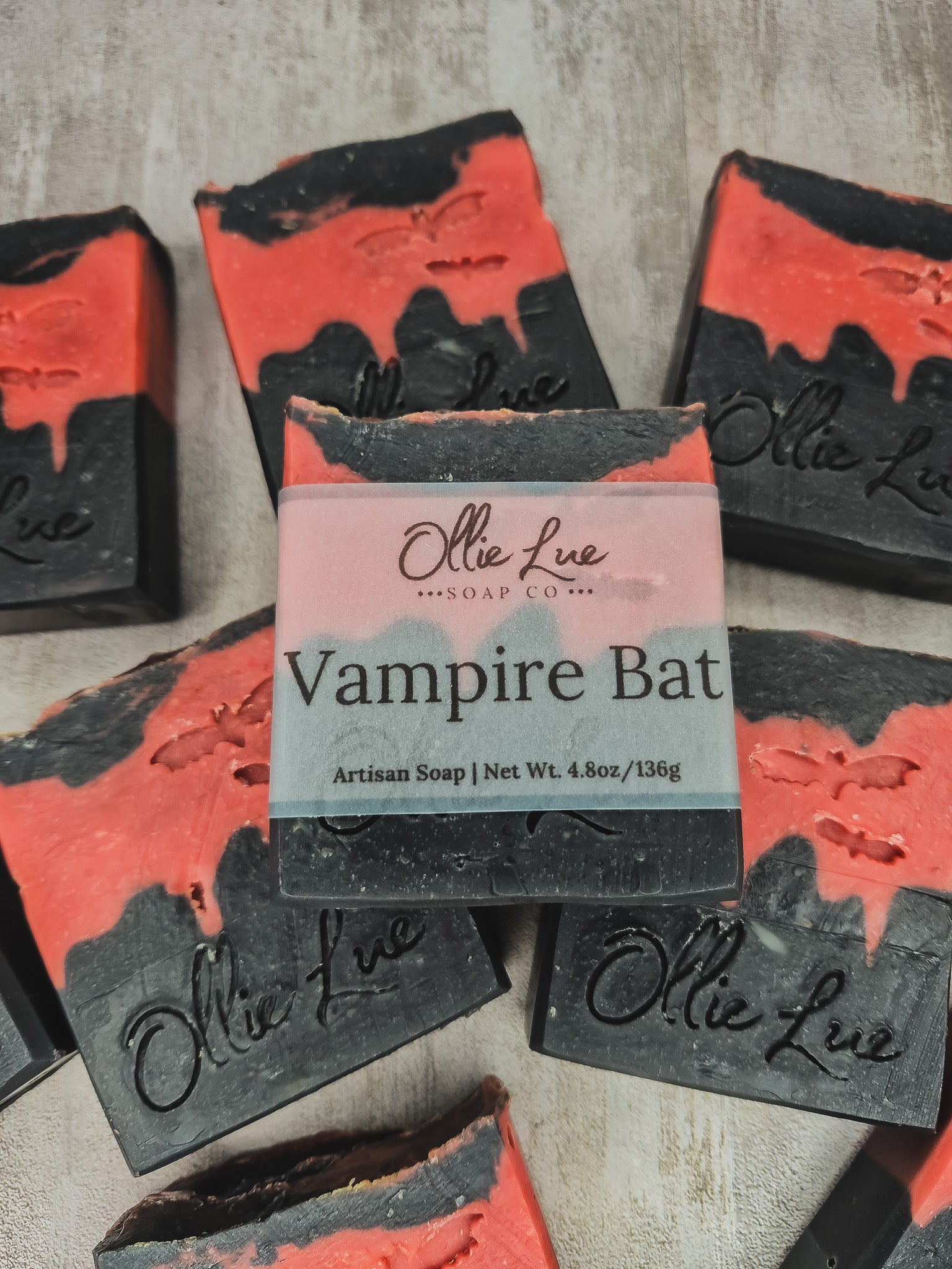 A bar of Ollie Lue Vampire Bat artisan soap is centered, surrounded by several identical soap bars arranged on a textured light gray surface. The soap has a black base with a bright red-orange marbled top layer and a pale pink label wrapped around the middle.
