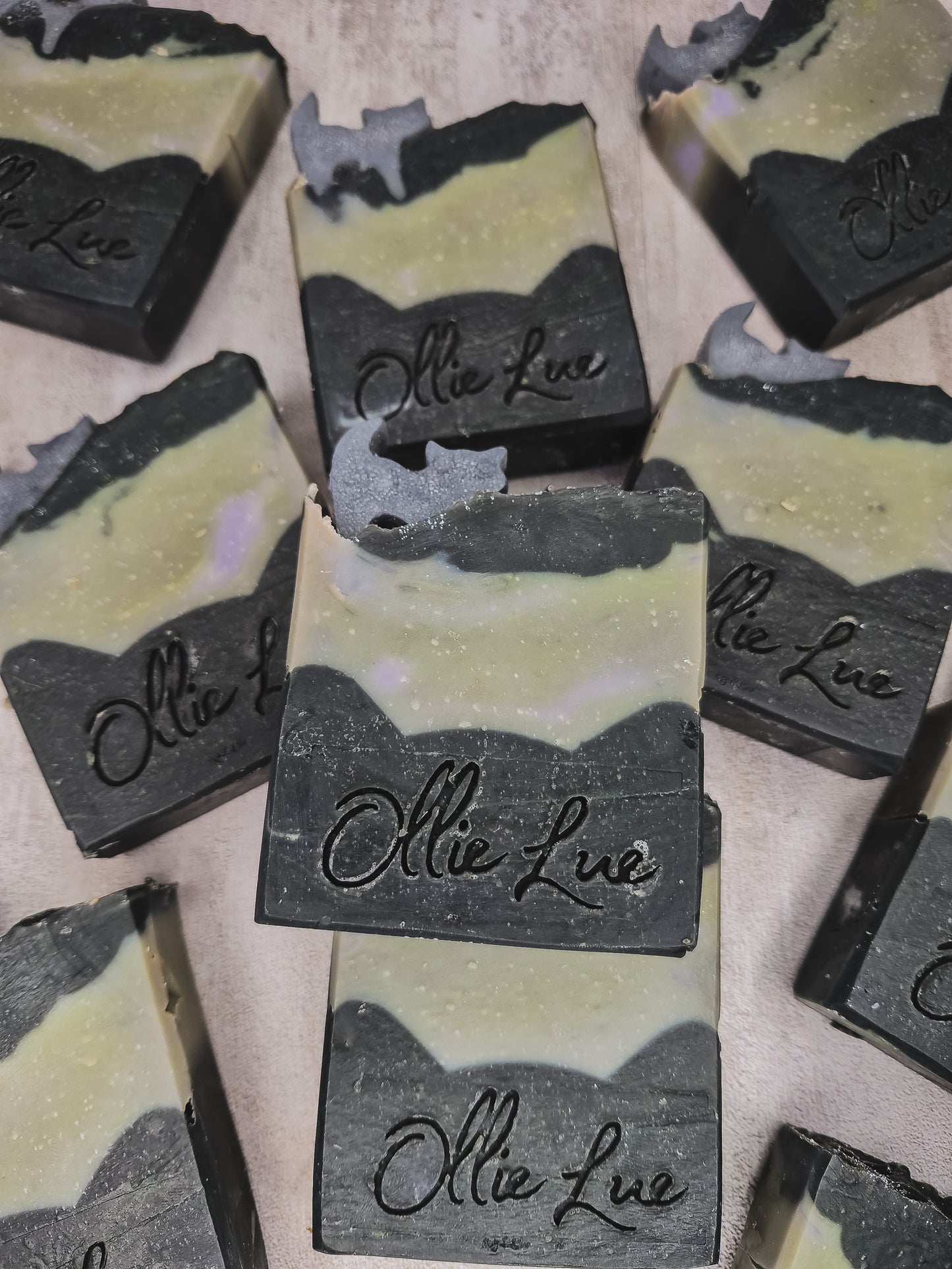 Several handmade soap bars with a marbled black and pale yellow-green design, featuring the brand name 'Ollie Lue' embossed on the front, arranged on a neutral surface.
