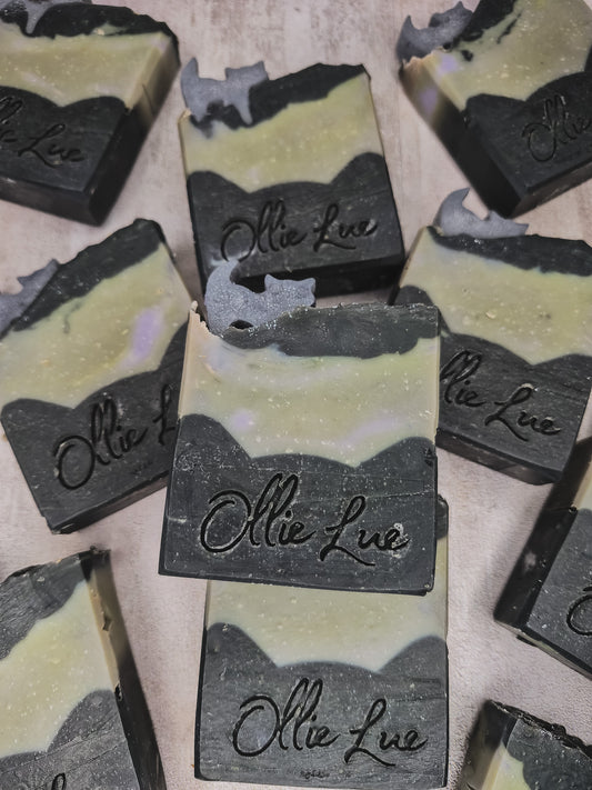 Several handmade soap bars with a marbled black and pale yellow-green design, featuring the brand name 'Ollie Lue' embossed on the front, arranged on a neutral surface.