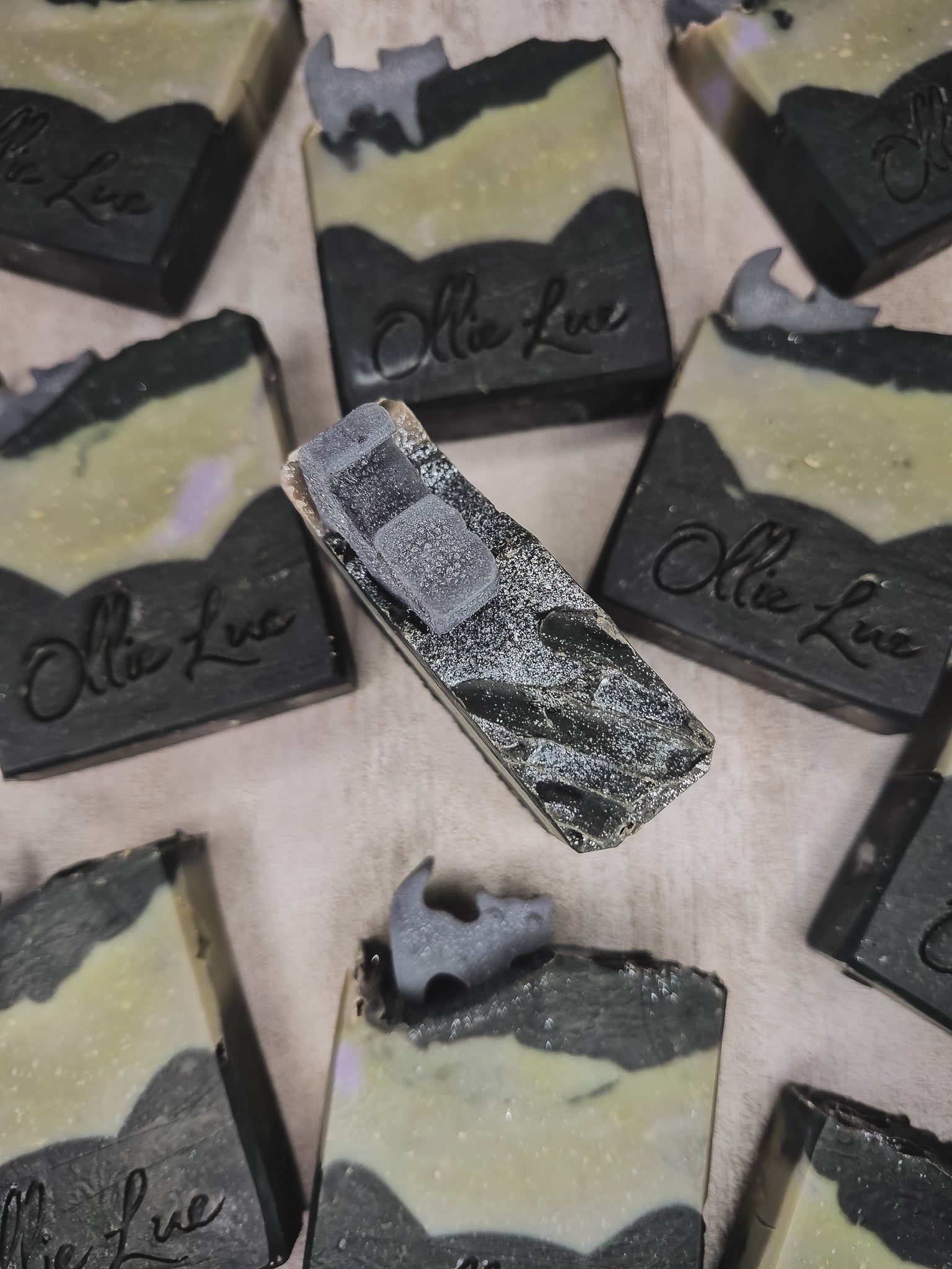 Several rectangular bars of handmade soap with a black and pale yellow marbled pattern, some showing rough, unpolished edges. One soap bar in the center is brushed with silver glitter.