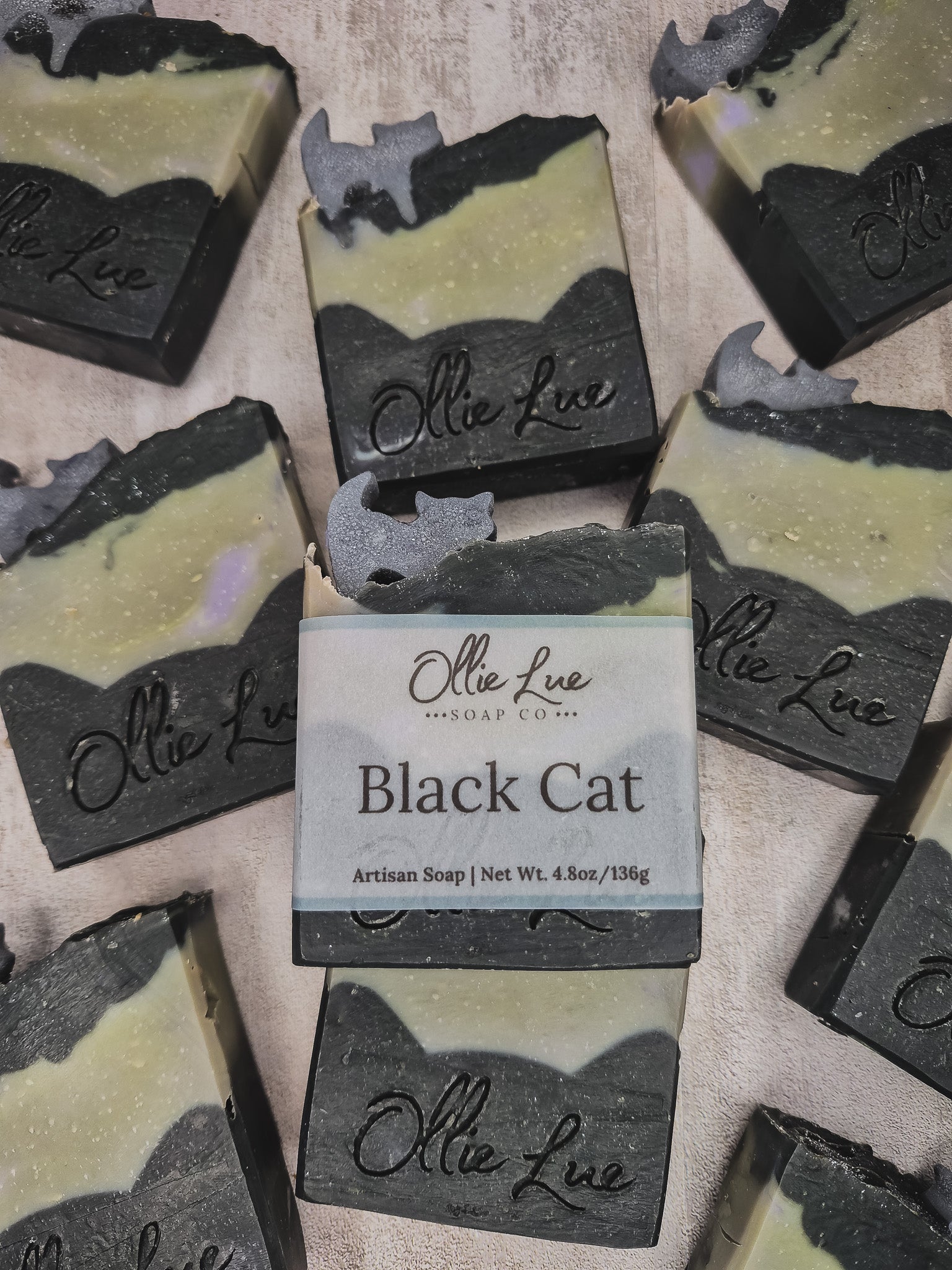 Several handmade soap bars are arranged on a light fabric background, with one central bar wrapped in a paper label that reads 'Olie Lue Soap Co