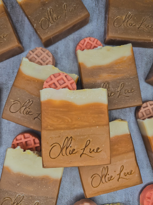 Several handmade soap bars with a layered tan, brown, and cream design are arranged on a textured gray-blue surface. The center soap bar is clearly engraved with the brand name 'Ollie Lue.
