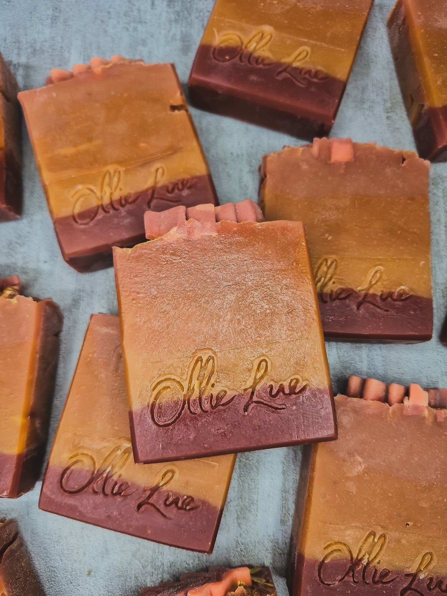Several handmade rectangular bars of soap with a warm gradient from golden orange to deep reddish-brown, each embossed with the brand name 'Olie Lue', arranged on a textured light blue-gray surface.