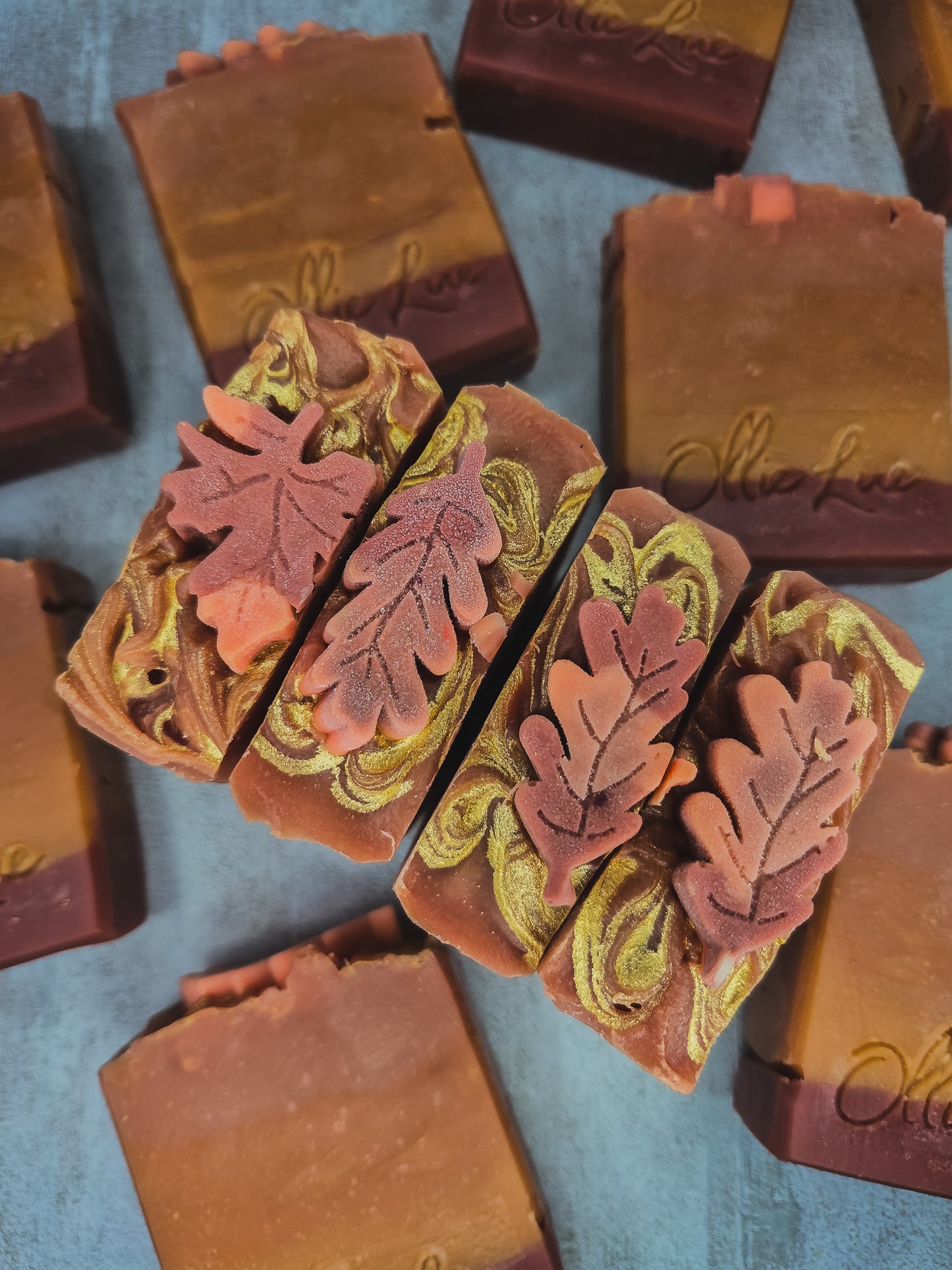 Handcrafted artisanal soap bars with autumn leaf designs, featuring marbled brown, terracotta, and olive green swirls, arranged on a textured blue-gray surface.