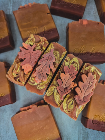 Handcrafted artisanal soap bars with autumn leaf designs, featuring marbled brown, terracotta, and olive green swirls, arranged on a textured blue-gray surface.