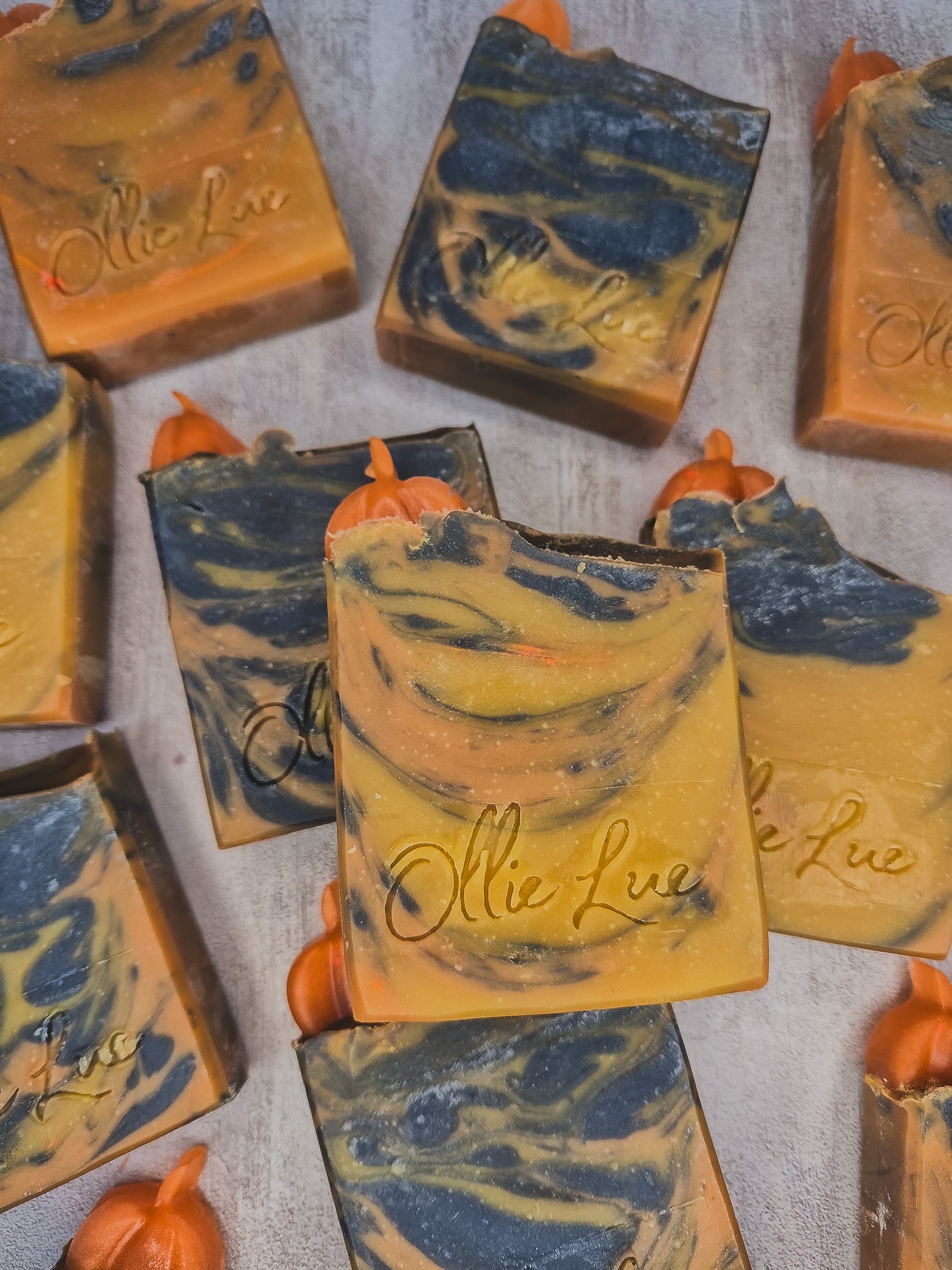 Multiple handmade rectangular bars of soap with orange and navy blue marbled swirls, each topped with a small orange pumpkin accent, arranged on a light gray textured surface. The soap has the brand name 'Olie Lué' embossed on the front.