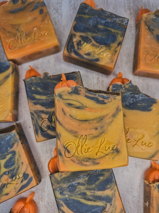 Multiple handmade rectangular bars of soap with orange and navy blue marbled swirls, each topped with a small orange pumpkin accent, arranged on a light gray textured surface. The soap has the brand name 'Olie Lué' embossed on the front.