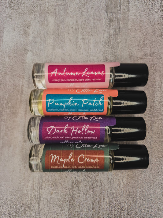 Four small roll-on perfume bottles labeled Autumn Leaves, Pumpkin Patch, Dark Hollow, and Maple Creme, arranged vertically on a textured gray background.