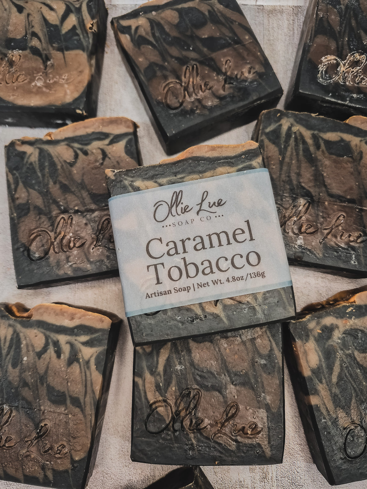 Several handmade soap bars are arranged on a light fabric background. The center soap is labeled 'Olle Lue Soap Co.