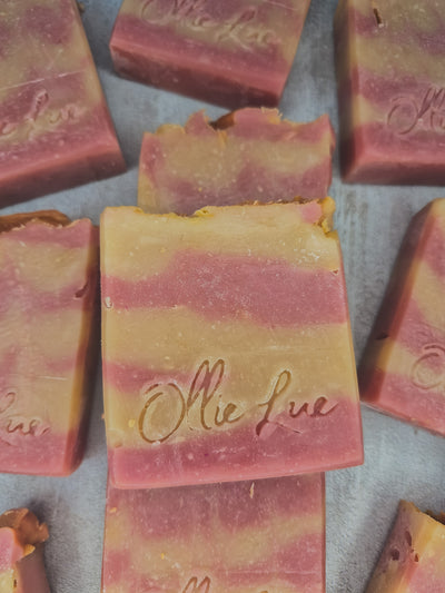 Several handmade rectangular bars of soap in pink and pale yellow marbled layers, arranged on a light gray surface. The center bar is upright and embossed with the brand name 'Ollie Lue.