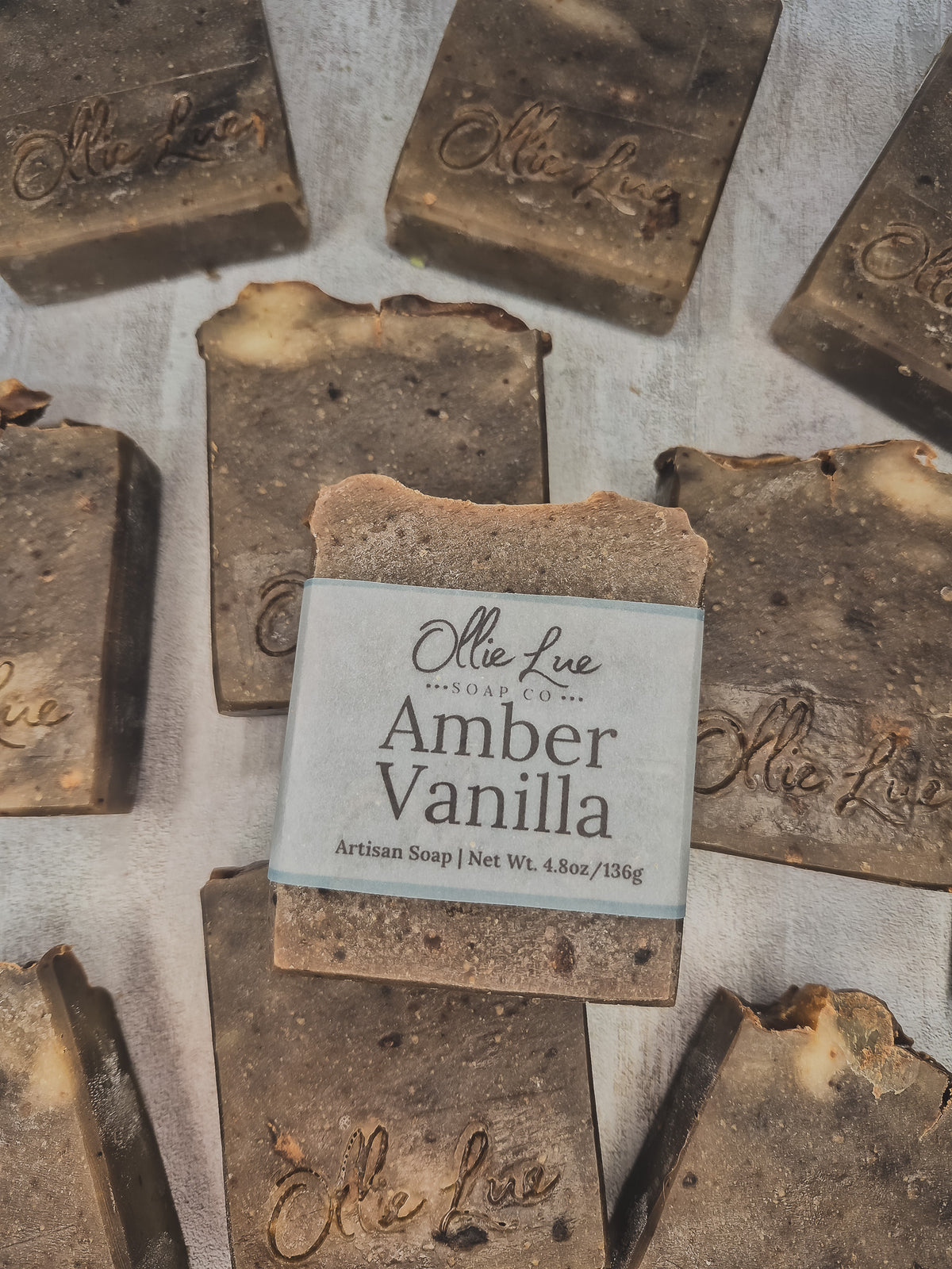 An artisan handmade soap bar labeled 'Amber Vanilla' by ollie lue soap co, shown in the center with other similar soaps arranged around it on a textured light background.
