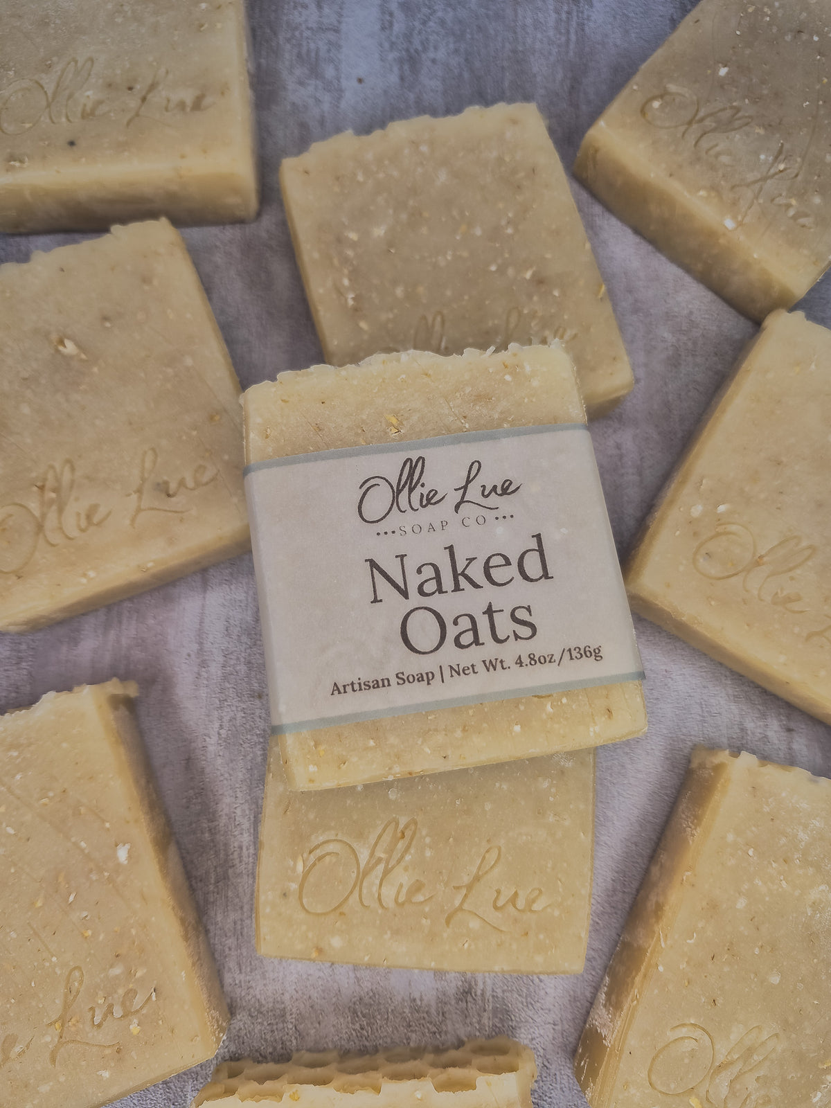 A bar of handmade soap wrapped in a beige paper label reading 'Olie LueSoap Co. Naked Oats Artisan Soap | Net Wt.