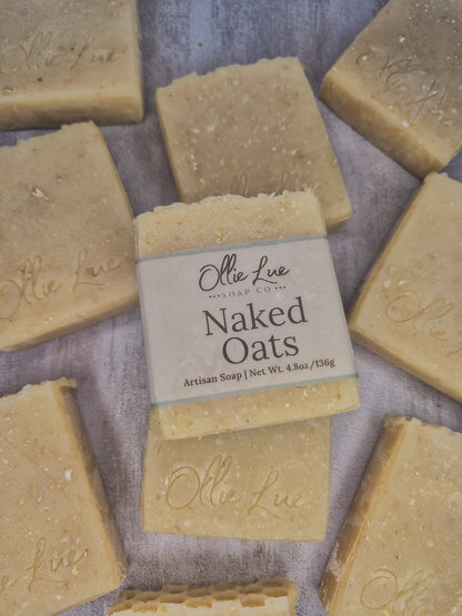 A bar of handmade soap wrapped in a beige paper label reading 'Olie LueSoap Co. Naked Oats Artisan Soap | Net Wt.