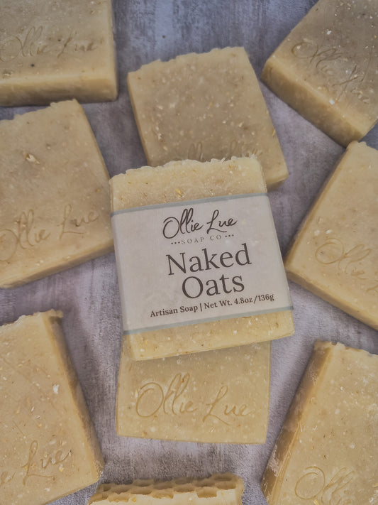A bar of handmade soap wrapped in a beige paper label reading 'Olie LueSoap Co. Naked Oats Artisan Soap | Net Wt.