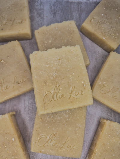 Several rectangular bars of natural soap in a warm beige-tan color, arranged on a textured gray surface. The soap bars are embossed with the brand name 'Ollie Lue'.