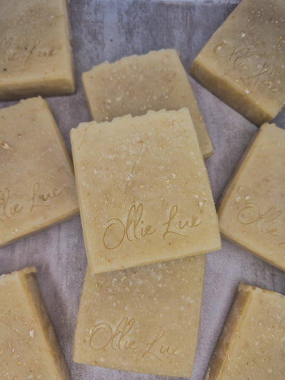 Several rectangular bars of natural soap in a warm beige-tan color, arranged on a textured gray surface. The soap bars are embossed with the brand name 'Ollie Lue'.