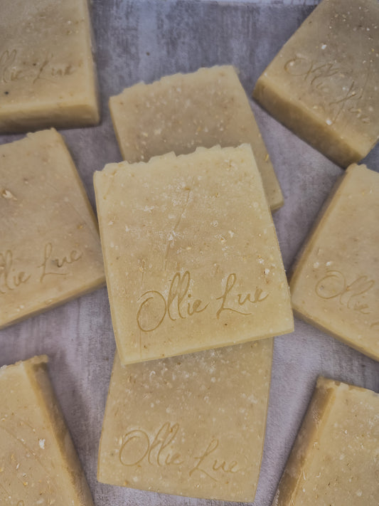 Several rectangular bars of natural soap in a warm beige-tan color, arranged on a textured gray surface. The soap bars are embossed with the brand name 'Ollie Lue'.