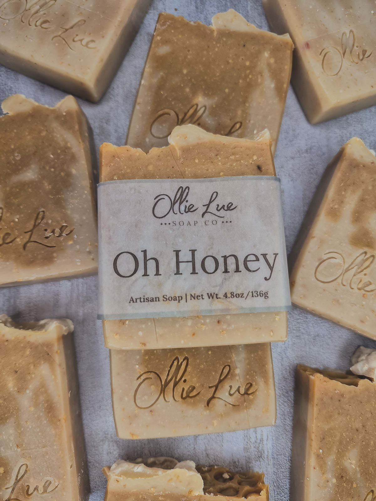 A stack of handmade artisan soap bars in a warm honey beige color, with a central bar wrapped in a paper label that reads 'Oh Honey' by 'Olie Lue Soap Co'.