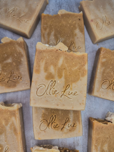Several handmade rectangular bars of soap in warm tan and beige tones are arranged on a light gray surface. The center bar is stacked on another, and the soap pieces are embossed with the brand name 'Ollie Lue'.