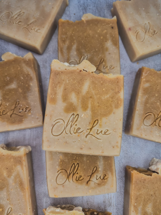 Several handmade rectangular bars of soap in warm tan and beige tones are arranged on a light gray surface. The center bar is stacked on another, and the soap pieces are embossed with the brand name 'Ollie Lue'.