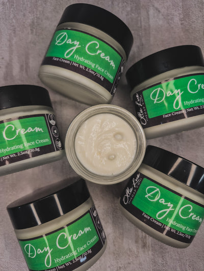 Five jars of 'Day Cream Hydrating Face Cream' arranged in a circle, with one open jar in the center showing the thick off-white cream. The jars have black lids and labels with green and white branding, set against a textured gray background.