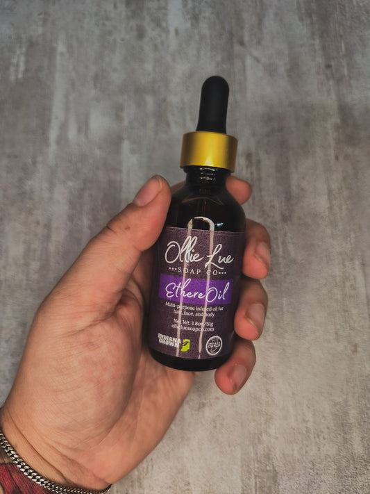 A hand holding a dark amber glass dropper bottle of Olive Lu Apothecary Ethereal Oil against a textured gray concrete background.
