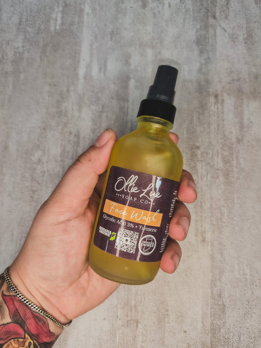 A hand holding a bottle of Ollie Lue Facial Wash against a textured gray concrete background.