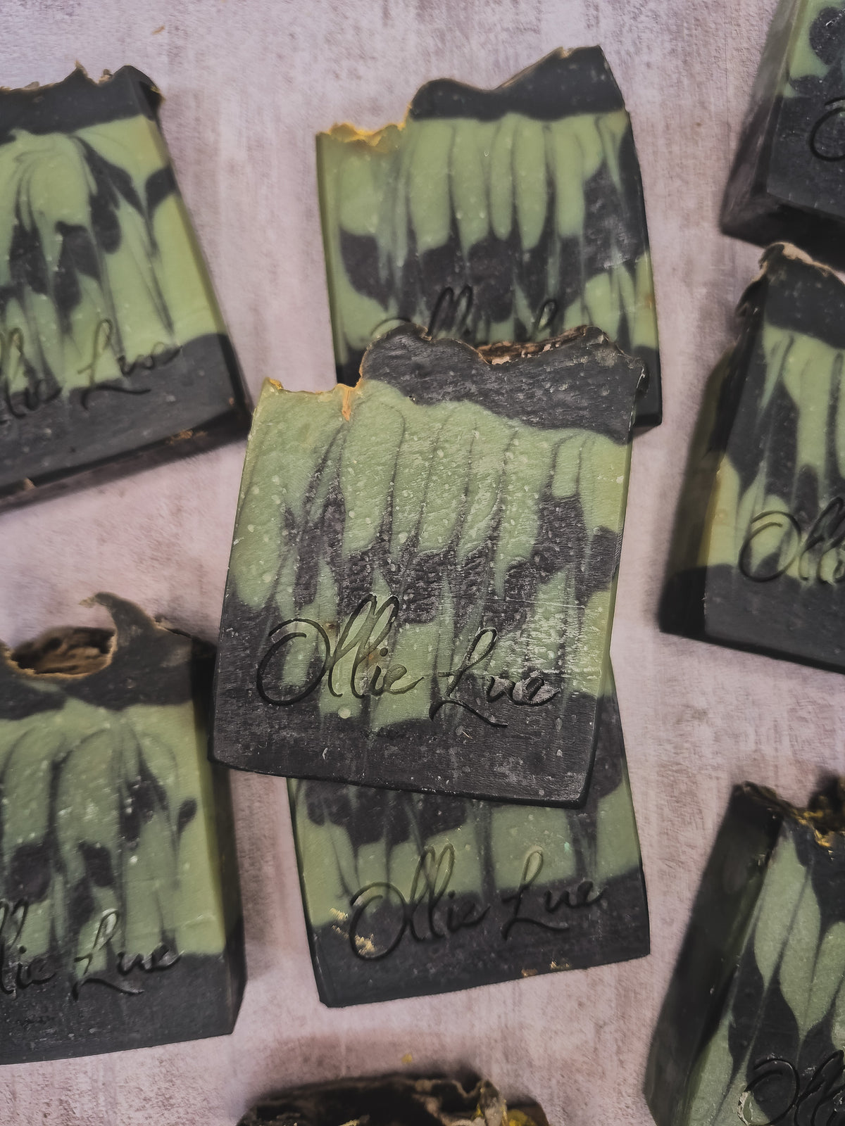 Several handmade soap bars with a green and black marbled pattern are arranged on a textured light gray surface. The center soap bar is upright and features the brand name 'Olie Lue' embossed in black script.