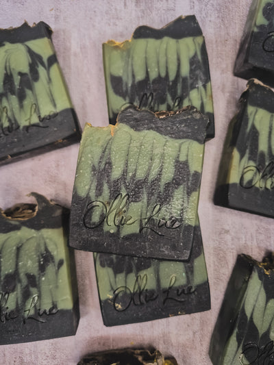 Several handmade soap bars with a green and black marbled pattern are arranged on a textured light gray surface. The center soap bar is upright and features the brand name 'Olie Lue' embossed in black script.