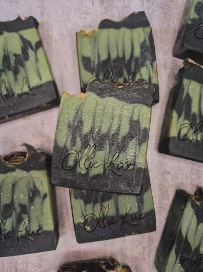 Several handmade soap bars with a green and black marbled pattern are arranged on a textured light gray surface. The center soap bar is upright and features the brand name 'Olie Lue' embossed in black script.