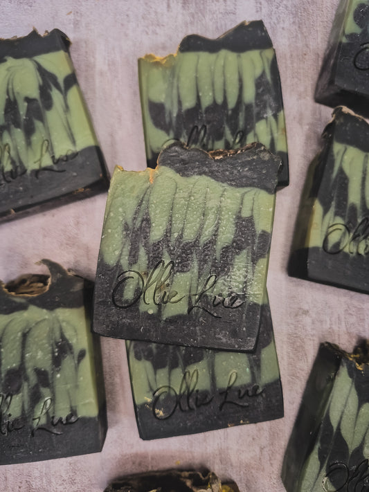 Several handmade soap bars with a green and black marbled pattern are arranged on a textured light gray surface. The center soap bar is upright and features the brand name 'Olie Lue' embossed in black script.