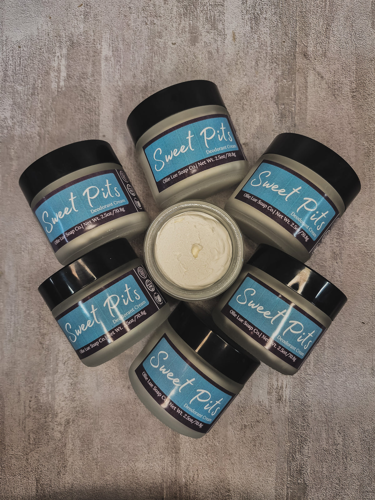 Six jars of Sweet Pits arranged in a circle on a textured gray surface, with one open jar in the center showing the creamy off-white oil.